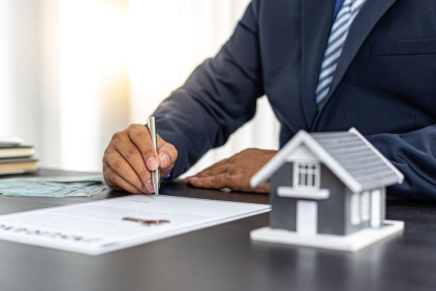 Un homme en costume signe un contrat officiel à côté d'une maquette de maison pour officialiser un compromis de vente immobilier.
